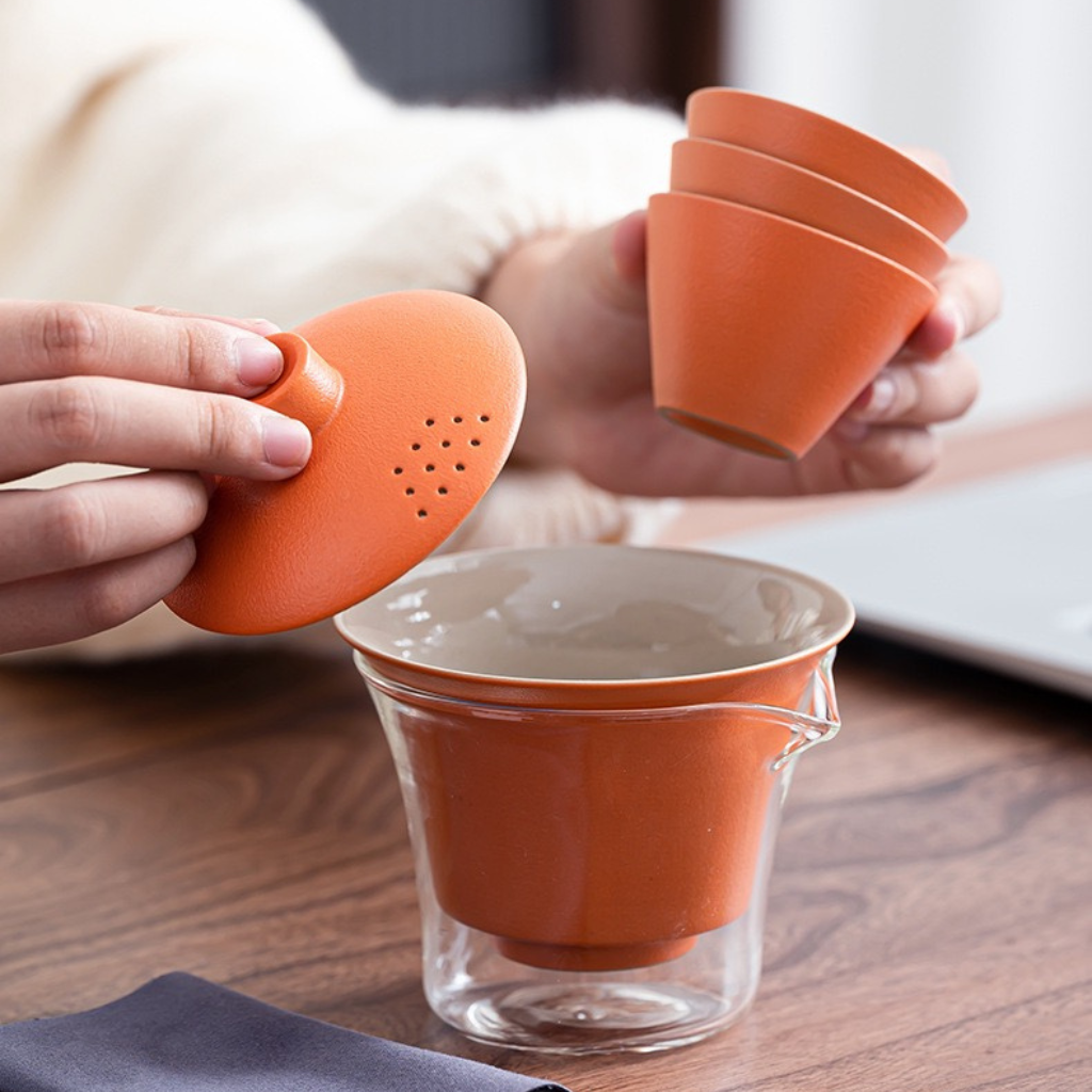 Person using an orange tea infuser with a glass mug on a wooden table.
