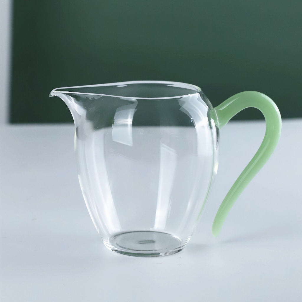 Minimalist transparent borosilicate glass Gongdao Bei tea pitcher with pastel green curved handle — elegant heat-resistant fair cup for Gongfu Cha and Chinese tea brewing rituals.
