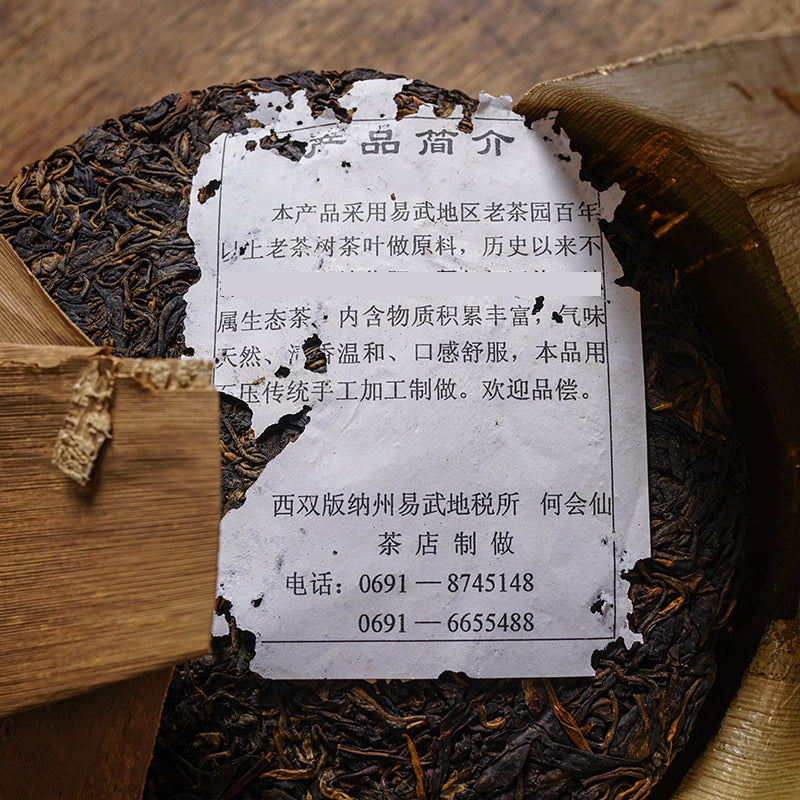 Vintage 2002 Huisheng Hao Yiwu Old Tea Garden Ancient Tree raw Pu-erh tea — aged Sheng Pu-erh 357g with partially unwrapped paper label and bamboo leaf wrapping, showing authentic dry-aged tea leaves from Yunnan – Tea & Leaf